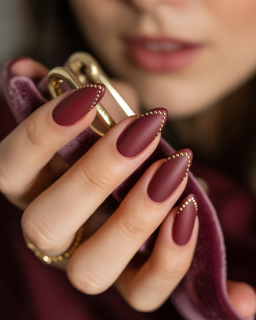 Almond-shaped nails with matte burgundy and chevron gold-studded tips.