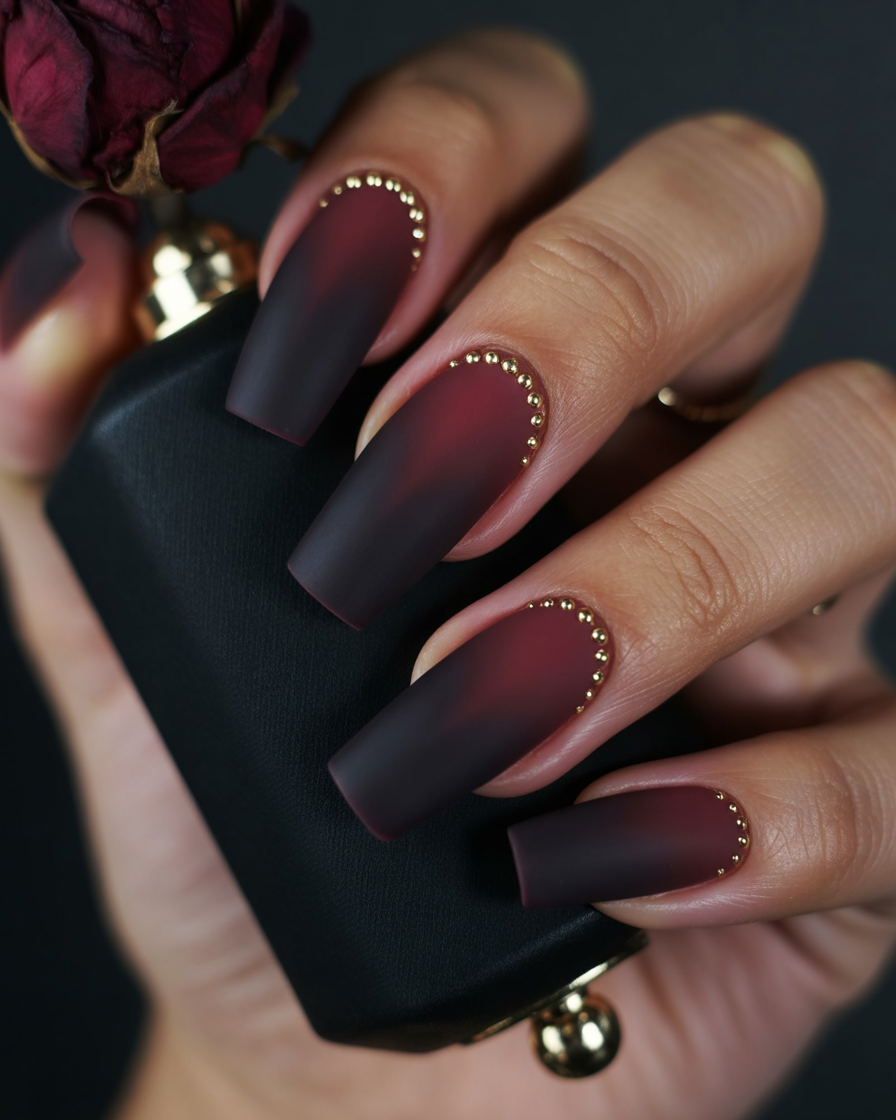 Ombre burgundy-black nails with gold-studded chevron detailing.