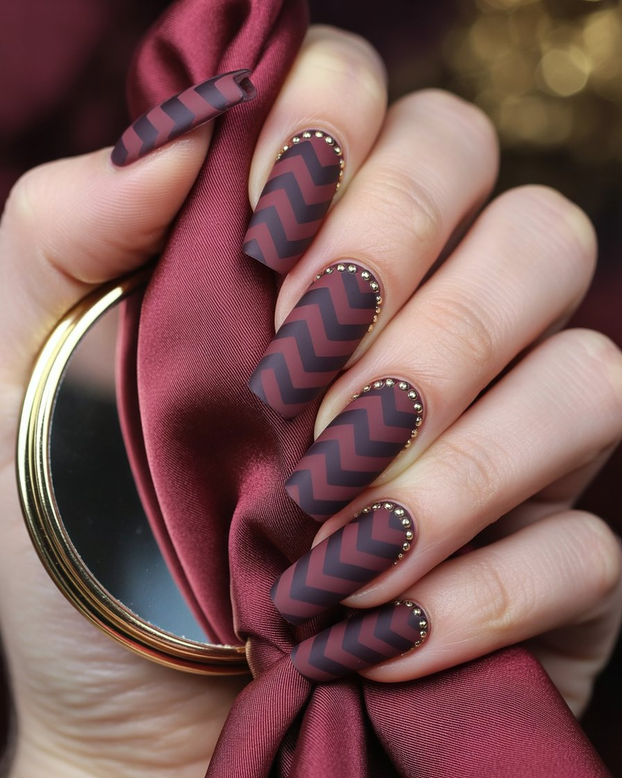 Nails covered in repeating matte burgundy chevrons with gold cuticle studs.