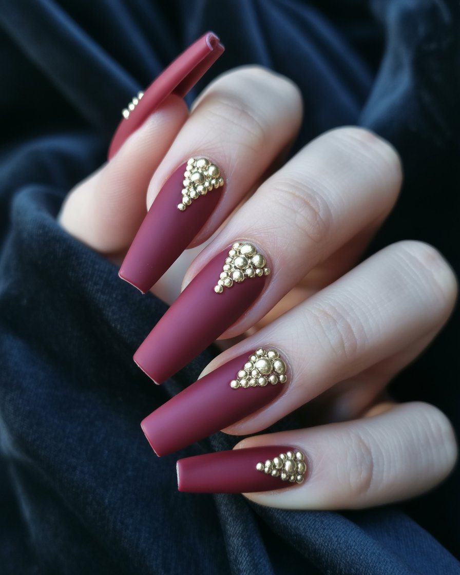 : Reverse chevron nails in matte burgundy with gold studs at the tip.