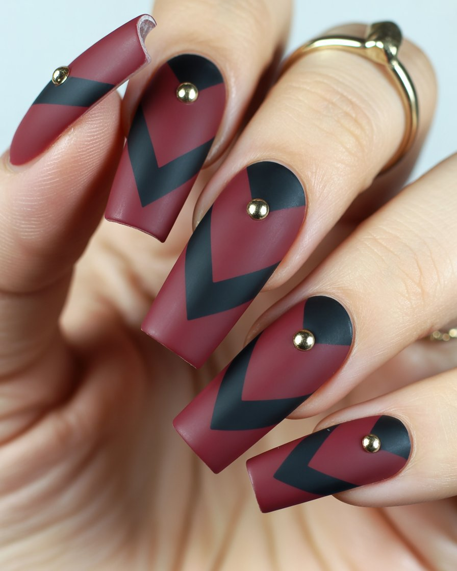 Matte burgundy nails with horizontal chevron stripes and central gold studs.