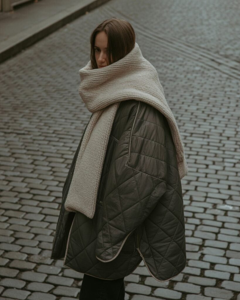 A person in a black quilted jacket and a large, chunky scarf on a moody street.