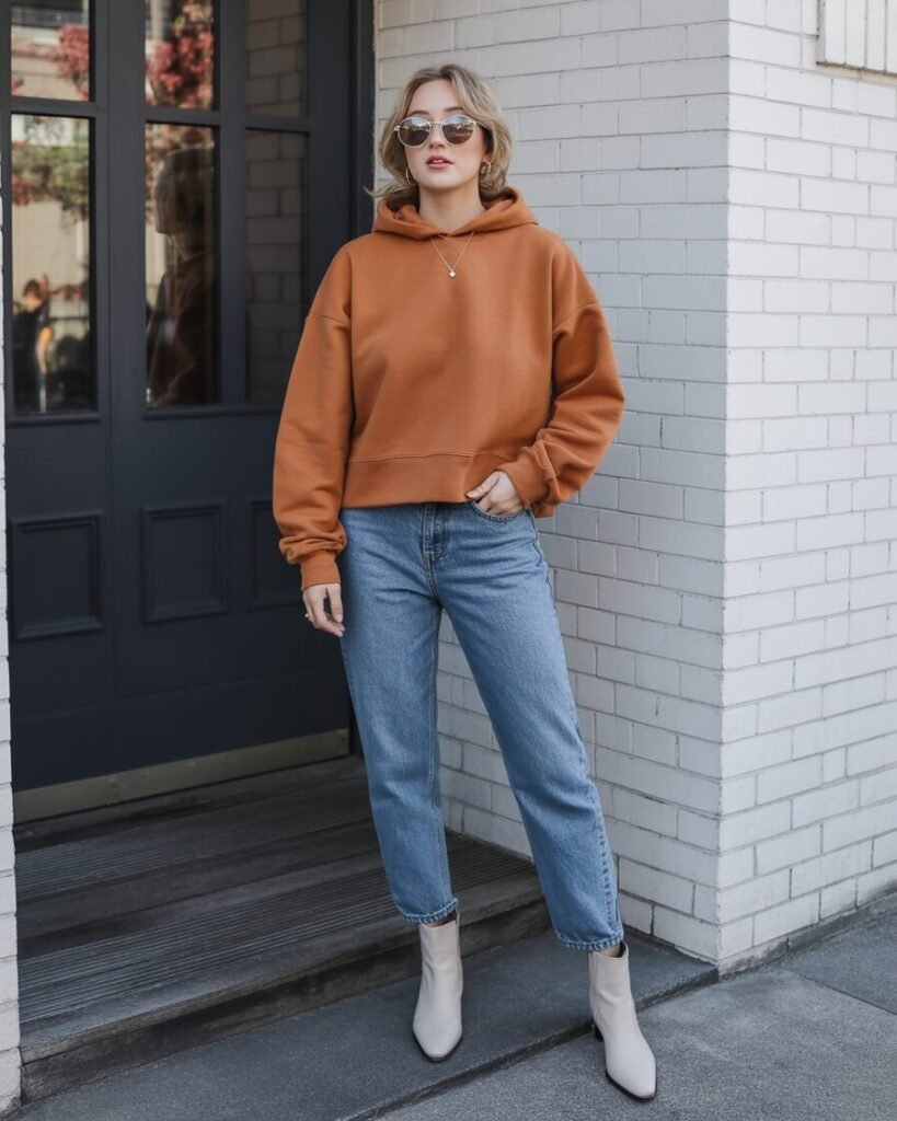 A woman wearing a structured, burgundy sweatshirt with straight-leg denim jeans and simple ankle boots, holding a coffee cup.